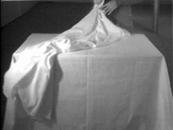 A person pulls a white cloth off a square table, with the cloth partly draped over the edge.