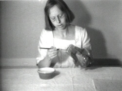 A woman sits at a table, dipping a brush into a bowl and painting a box.