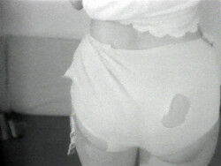 Person wearing torn shorts with visible holes, standing in a room with a bed in the background.