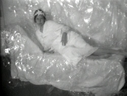 Person wearing a robe and head wrap lies on a couch covered in clear plastic sheets.