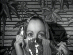 A person holds a shiny scarf over their mouth and nose, with wide eyes looking forward. Plants in the background.