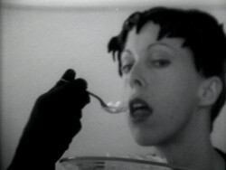Person with short dark hair is being fed with a spoon by someone wearing a black glove.