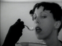 Person with short dark hair is being fed with a spoon by someone wearing a black glove.