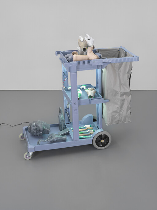 A blue utility cart holds mannequin body parts, brushes, and cleaning tools beside a grey laundry bag.
