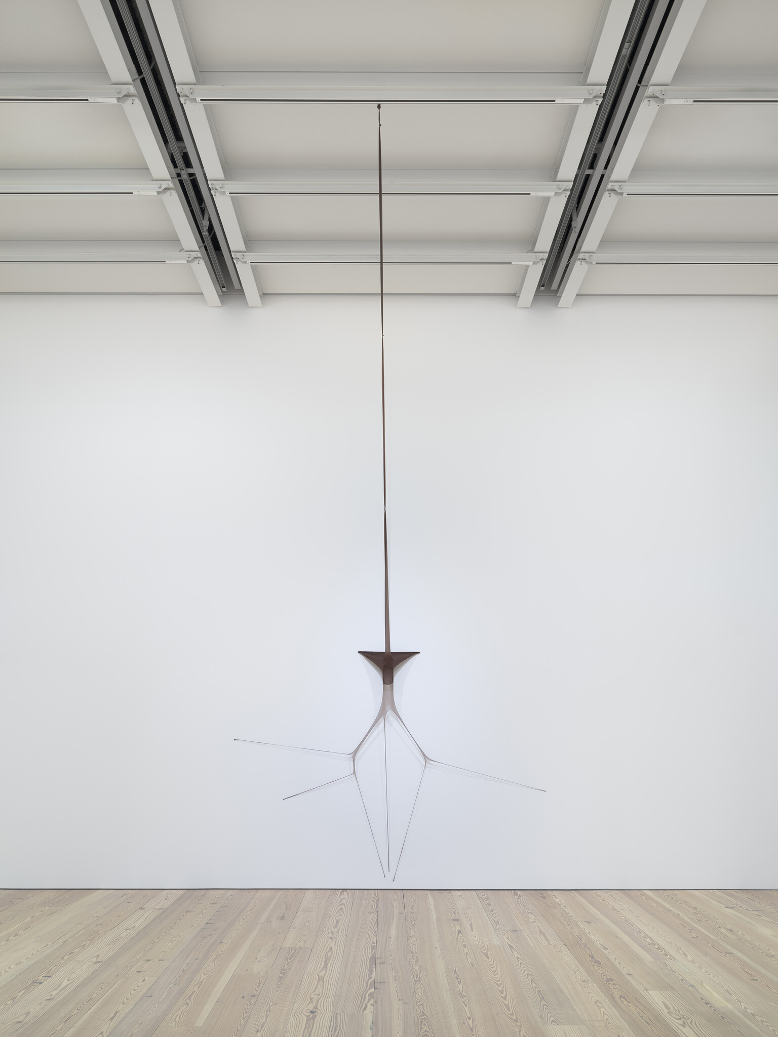 A slim, spider-like metal sculpture hangs from the ceiling to the gallery wall.