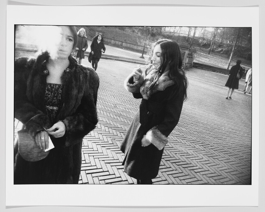 A black and white photograph capturing two women in the foreground, one exhaling smoke and the other lighting a cigarette, with other individuals walking in the background, all set against a herringbone-patterned pavement.