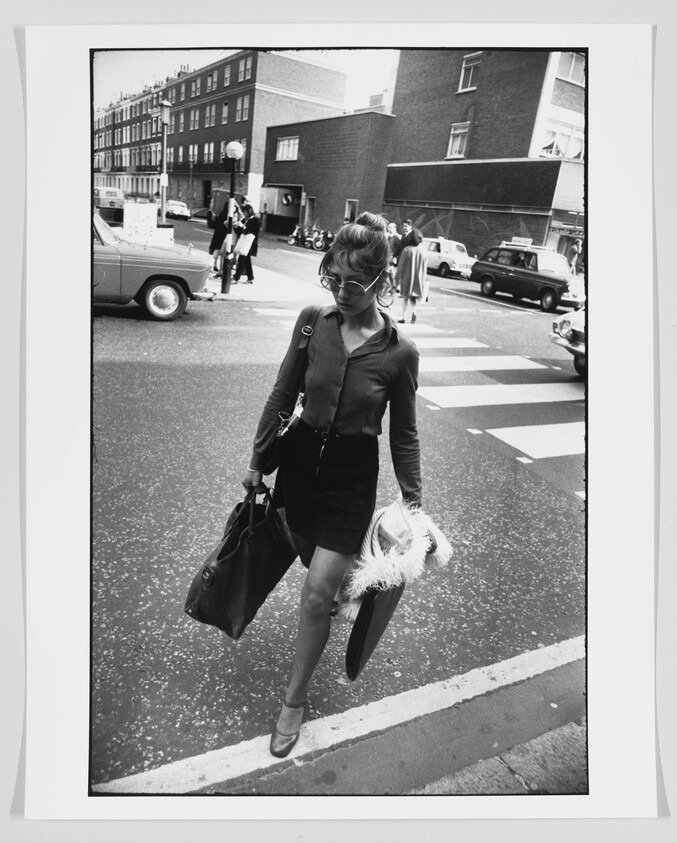 Young woman crosses a city street carrying two large bags and a fur-trimmed coat.