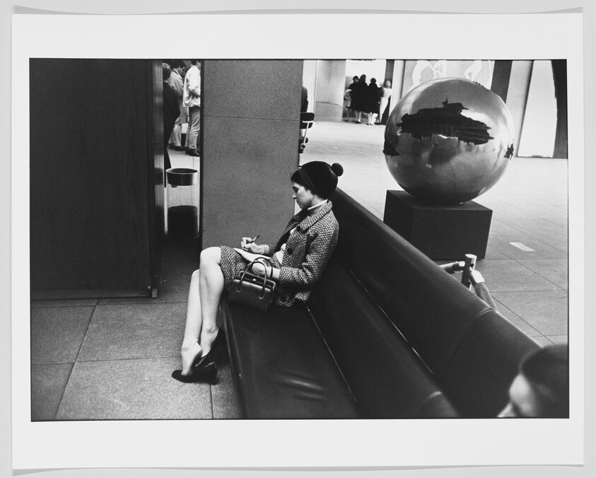 A woman in a hat sits on a bench writing in a notebook beside a reflective globe sculpture.