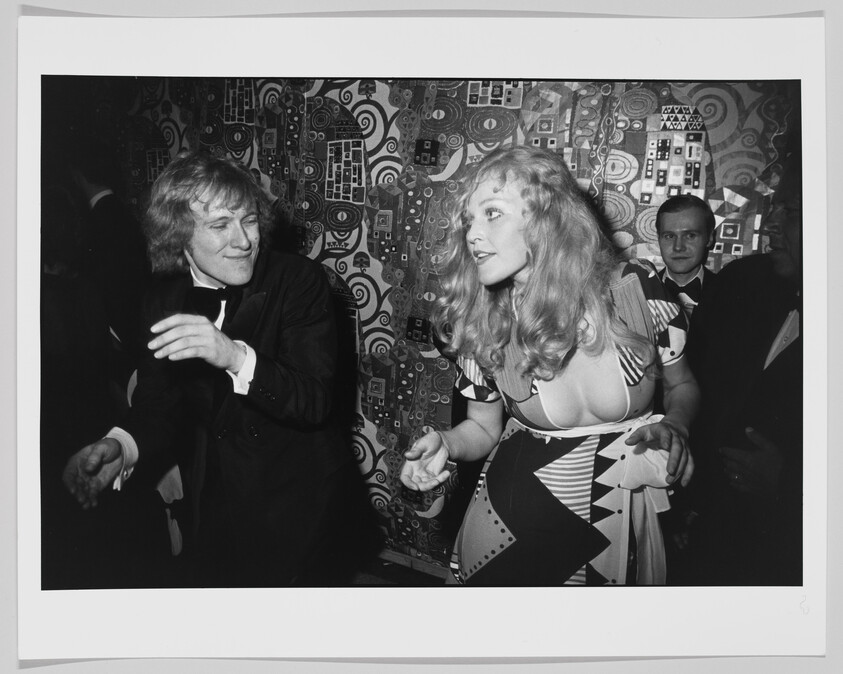 A smiling man and a woman in a patterned dress dance and lean toward each other.