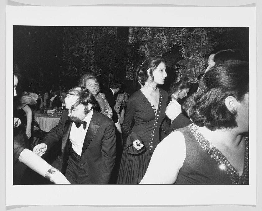 A group of well-dressed people dancing at a crowded formal party, woman in dark dress looking right.