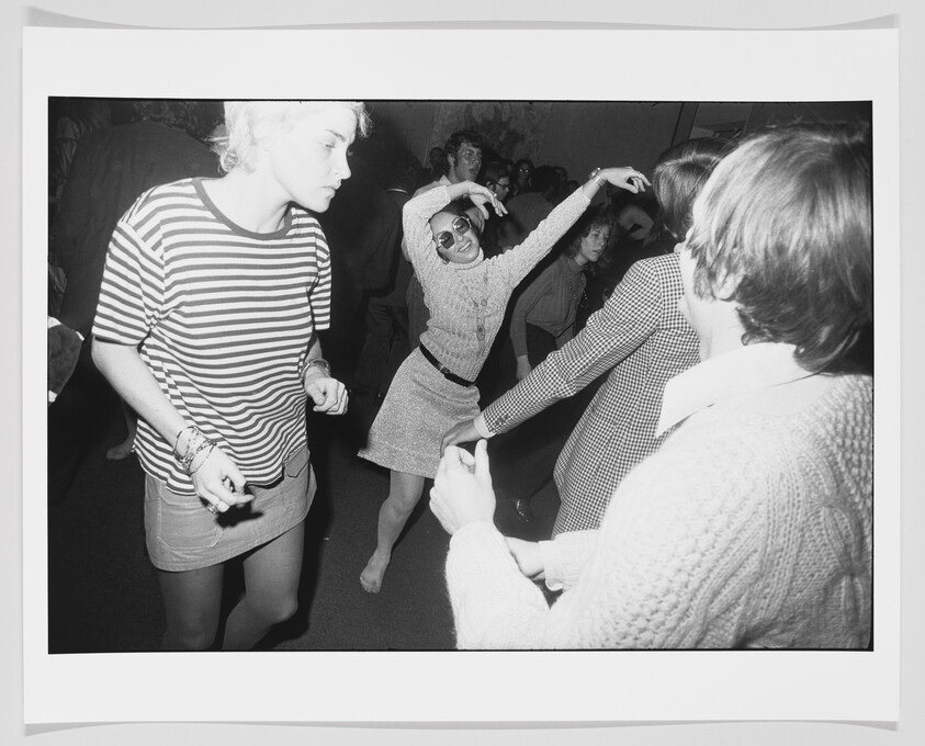 Young people dancing energetically in a crowded room, a woman in sunglasses striking a dramatic pose.