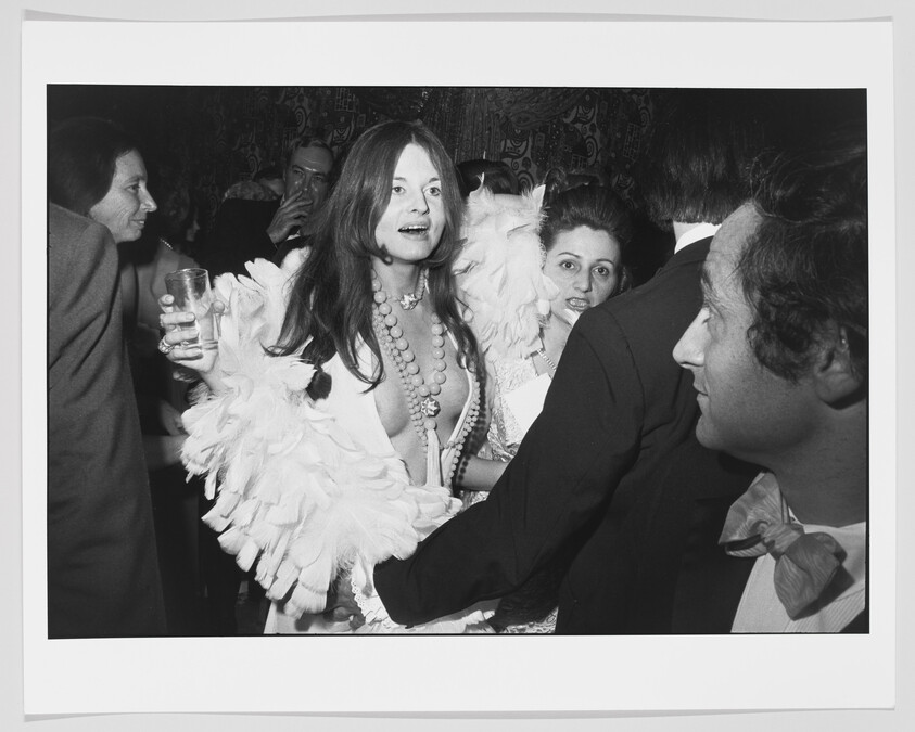 A woman wearing a feather boa and long beads holds a drink while talking in a crowded party.