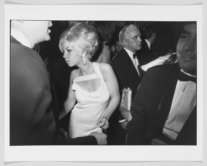 A woman in a white evening dress leans through a crowd at a formal party, holding a man's hand.