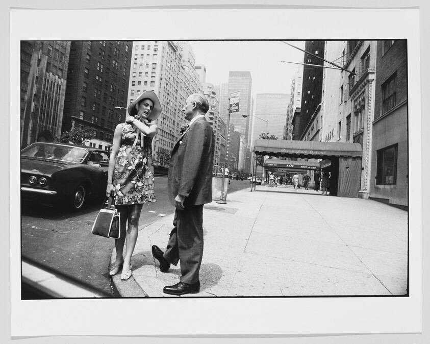 A woman in a dress and hat stands on a city sidewalk, talking to a man in a suit.