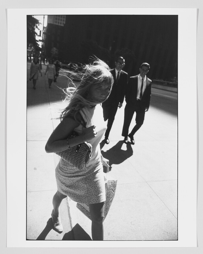 Young woman walks briskly down a sunny city sidewalk with her hair blowing and a handbag.