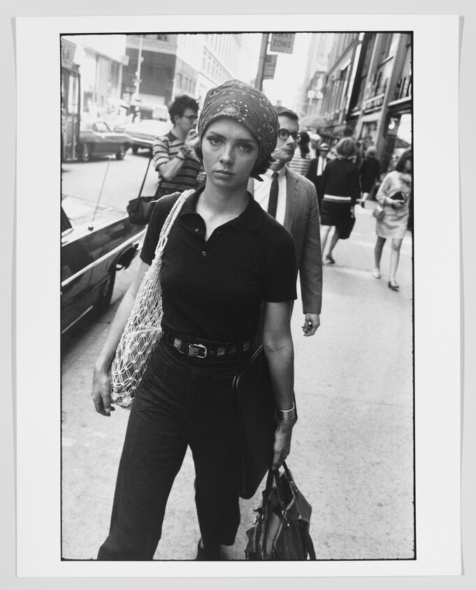 A black and white photograph of a young woman walking on a city street, looking directly at the camera with a neutral expression. She is wearing a dark shirt, pants, a patterned headscarf, and carries a net bag on her shoulder and a purse in her hand. Behind her, other pedestrians and vintage cars can be seen, suggesting the photo was taken in a previous decade.
