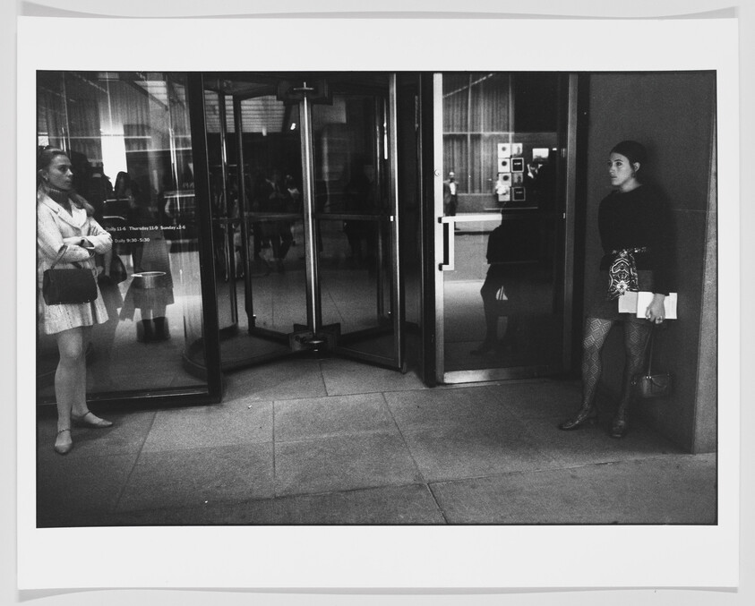 A black and white photograph capturing a moment outside a building with revolving doors. Two women stand on opposite sides of the entrance; one is exiting and looking to the side, while the other is entering, pausing and looking straight ahead. Reflections and interior details are visible through the glass doors, suggesting a busy indoor environment.