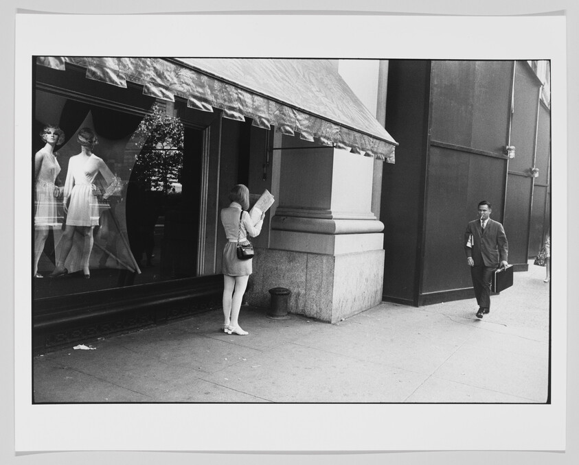 Woman stands reading a map outside a storefront while a man in a suit walks toward her.