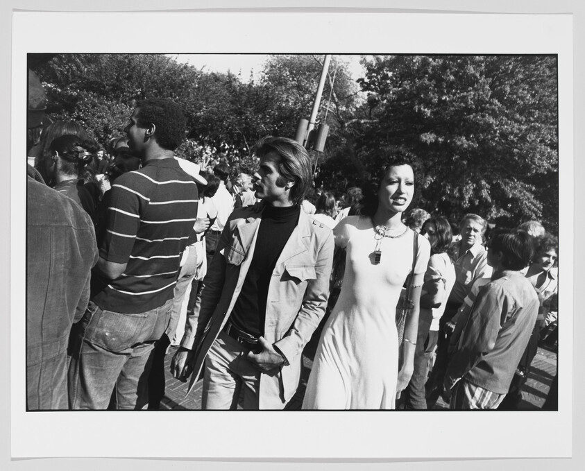 A black and white photograph capturing a diverse crowd of people, possibly at an outdoor event or gathering. In the foreground, a woman in a white dress stands out, looking to the side with a smile, while various individuals around her are engaged in conversations or looking in different directions. The setting appears to be a sunny day with trees in the background.