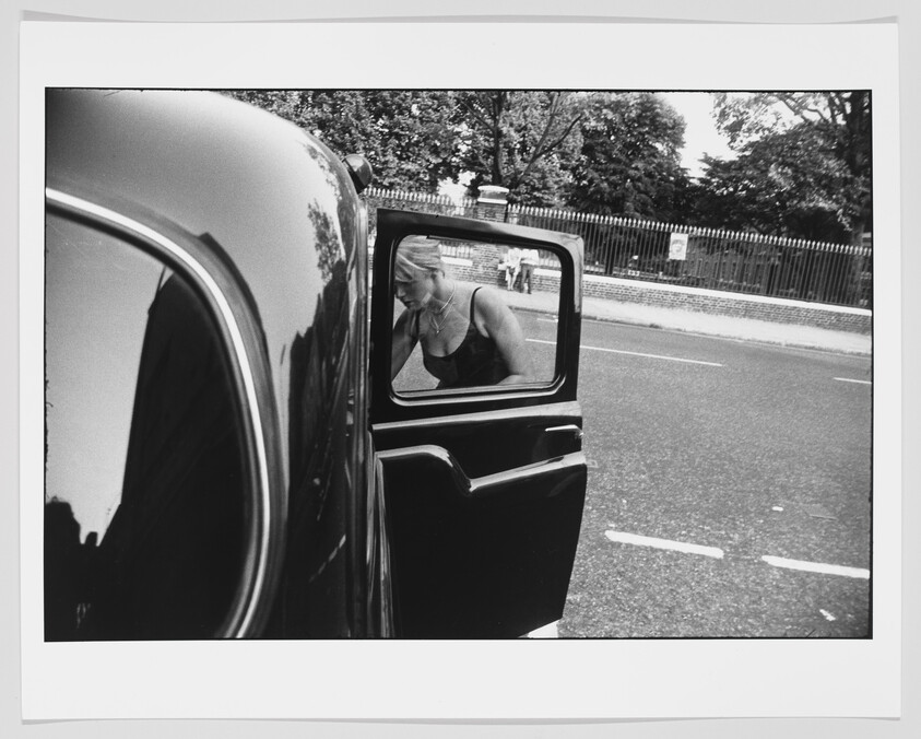 A black and white photograph capturing the reflection of a woman in a car's side mirror. The car door is open, and the woman appears to be seated inside, looking down. The background shows a street scene with a fence and trees.