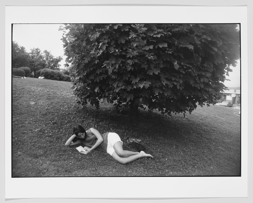 A black and white photo of a person lying on the grass under the shade of a large tree, reading a book. The setting appears to be a park with a clear sky and some buildings in the background.