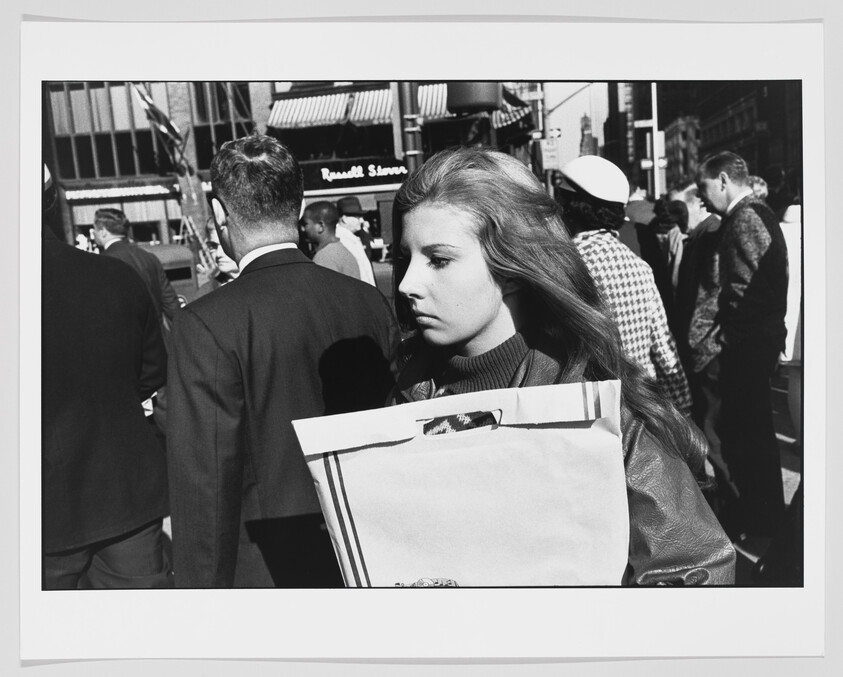 Young woman holds a large shopping bag while walking through a crowded city sidewalk.