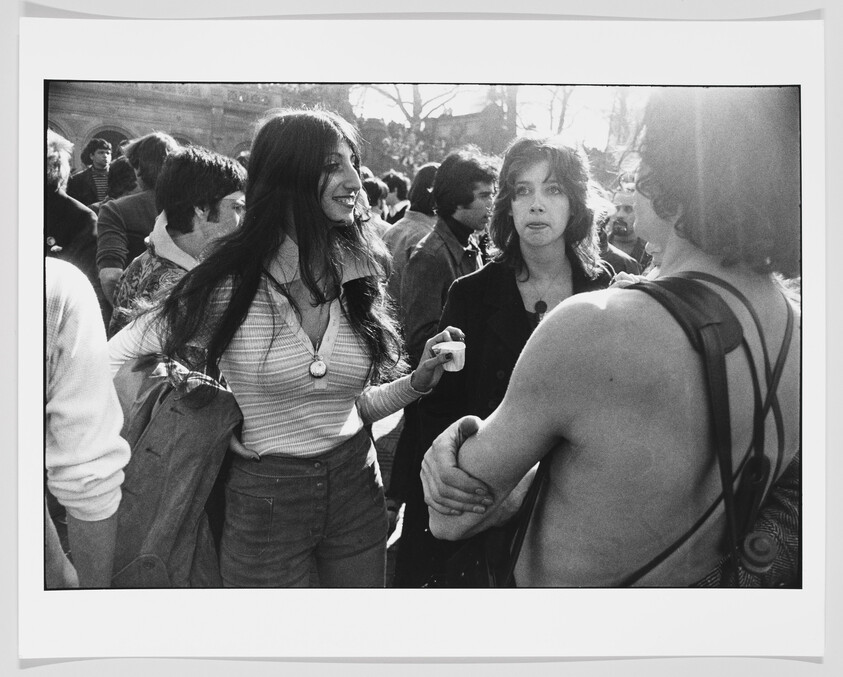 Young woman smiling and talking to a group in a crowded outdoor gathering while others listen.