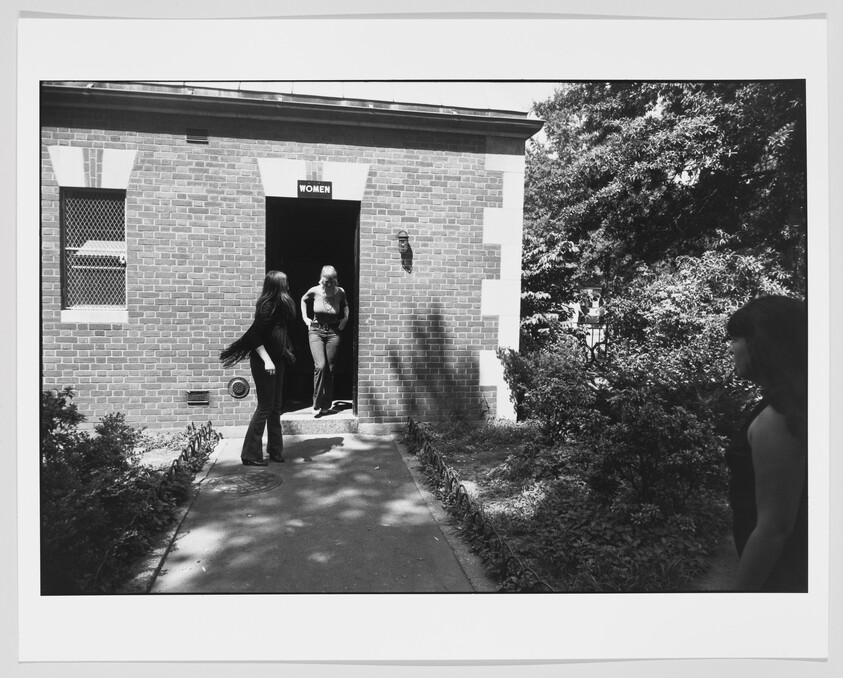 Three women near a brick restroom entrance labeled "WOMEN", one stepping out while others stand nearby.