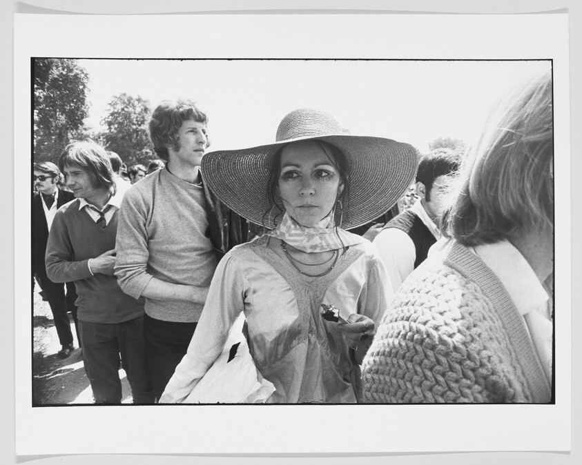 Young woman wearing a wide-brimmed hat stands in a crowded outdoor gathering, holding a small object.