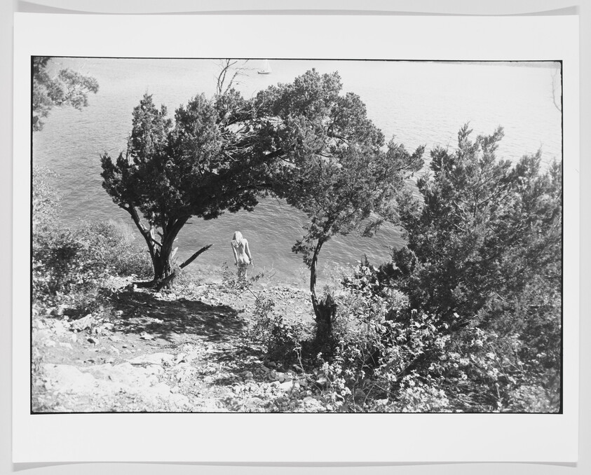 Black and white photograph of a scenic view showing a person standing by the water's edge under the shade of trees, with a glimpse of a sailboat in the distance on the water. The image is framed with a white border.