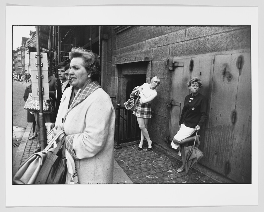 Women wait on a city sidewalk, one leaning against a building entrance while holding a bag.