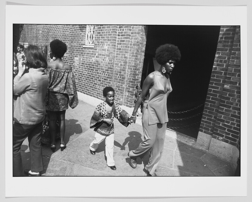 Woman with afro walks hand-in-hand with young boy past a brick building.