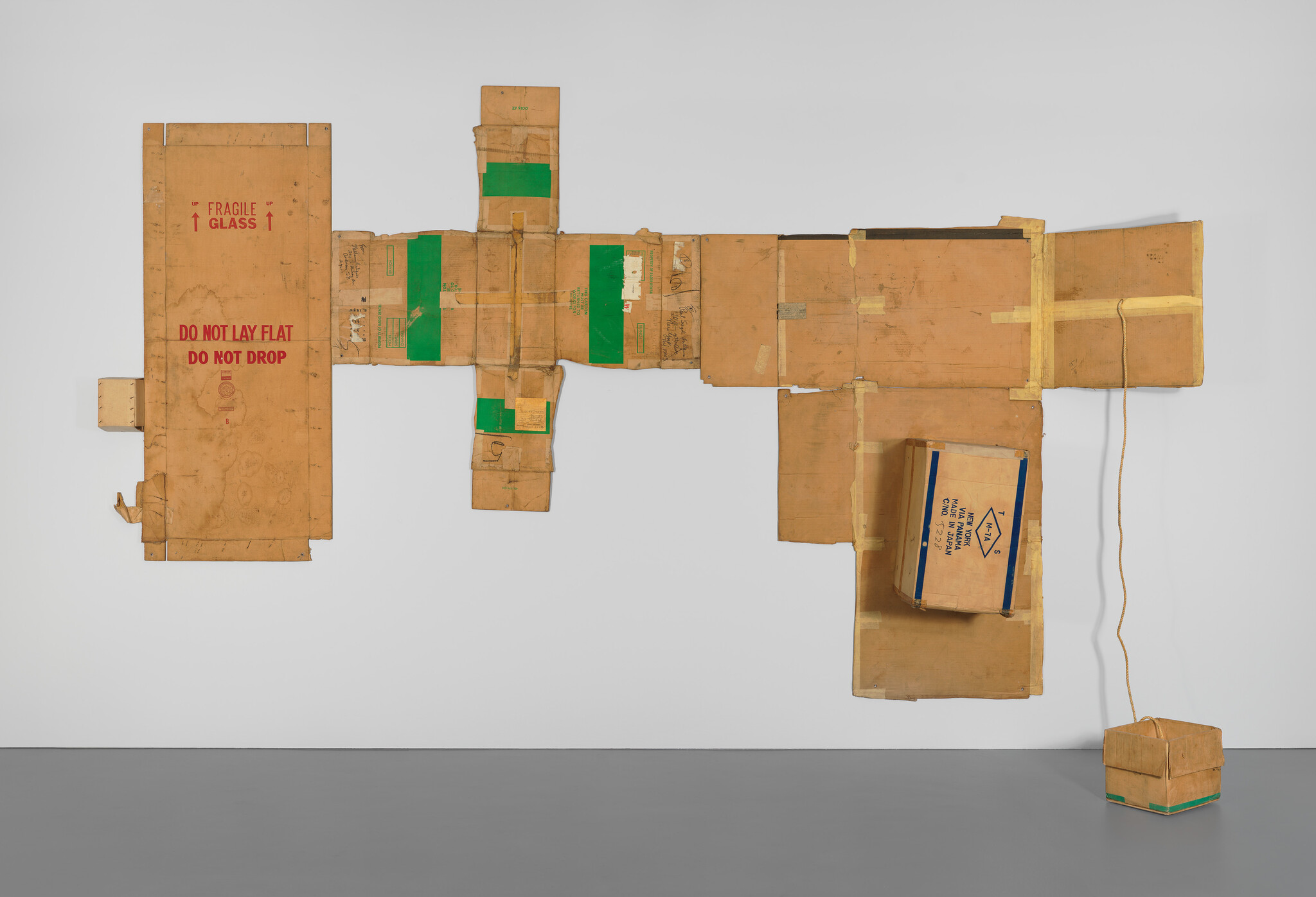 A large, abstract wall sculpture made from various sizes of cardboard boxes, some with shipping labels and tape, arranged in a cross-like formation and mounted on a white gallery wall. The sculpture features printed warnings such as "FRAGILE GLASS" and "DO NOT LAY FLAT DO NOT DROP," with a small cardboard box suspended by a string at the right end.