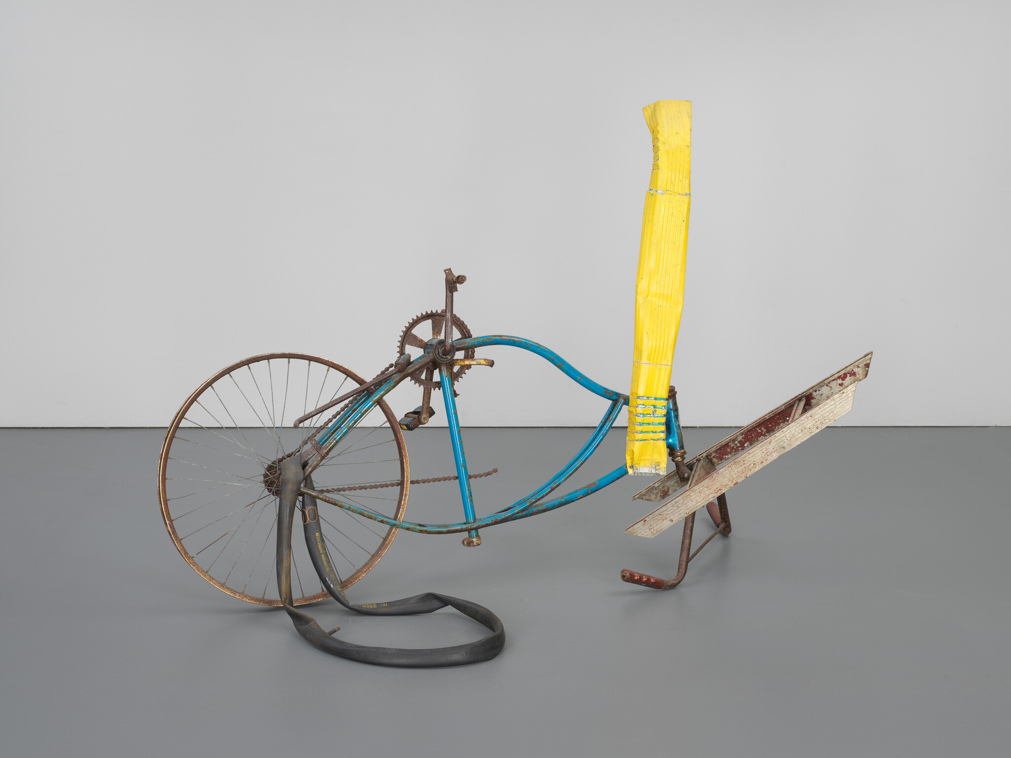 A sculpture composed of a disassembled bicycle with a blue frame, its parts rearranged and attached to a yellow-painted column and a brown ironing board, displayed on a grey floor against a white wall.