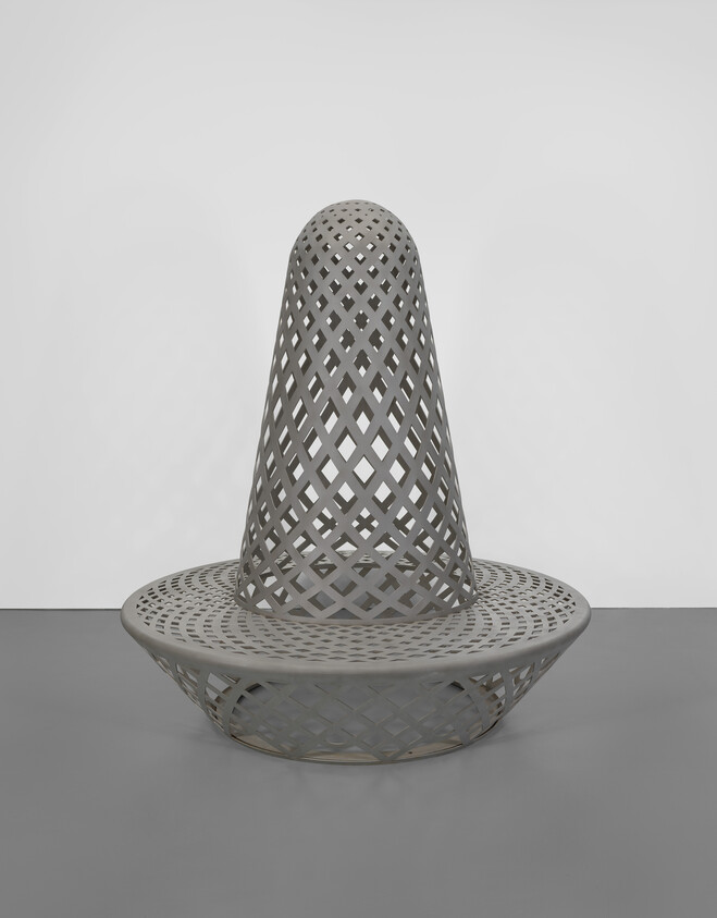 Large gray lattice metal sculpture with conical top and circular base on gallery floor.
