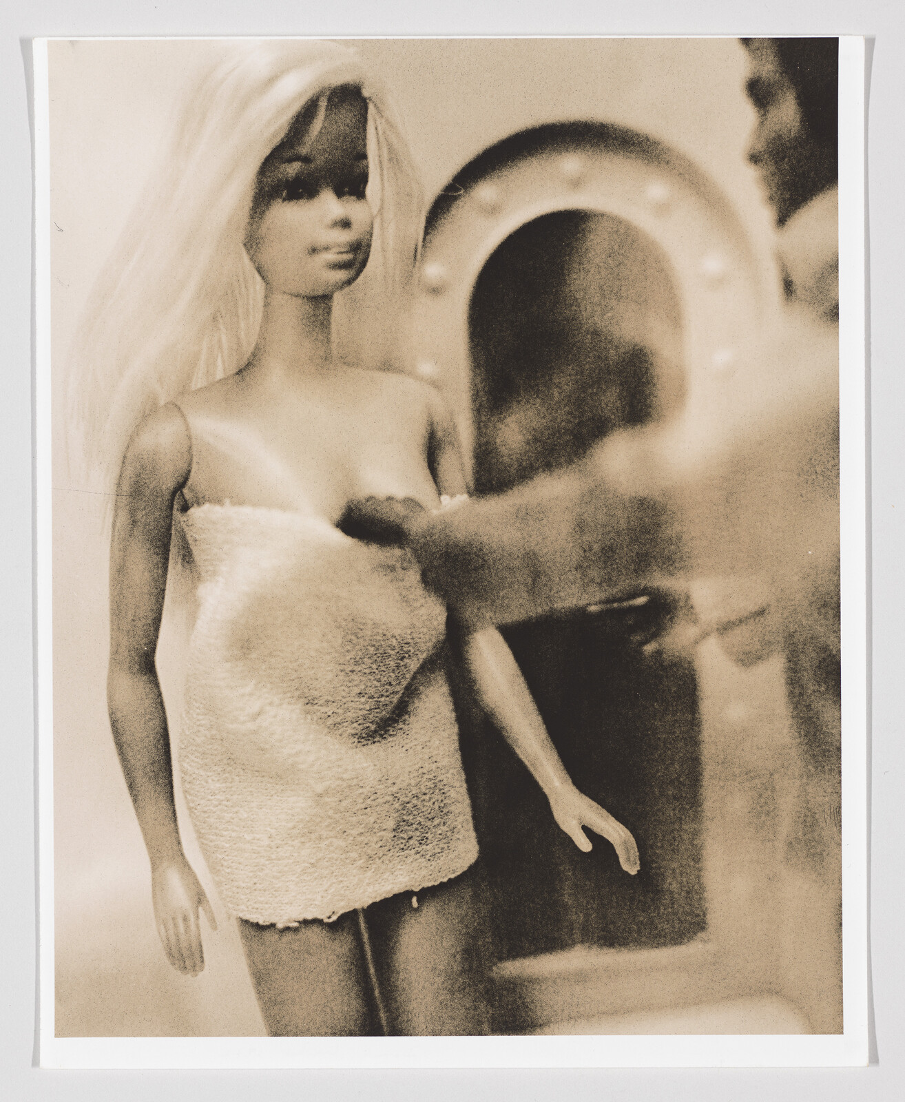 A vintage-looking photograph of a Barbie doll wrapped in a towel, standing next to a mirror that reflects its image. The photo has a sepia tone, adding to its retro aesthetic.