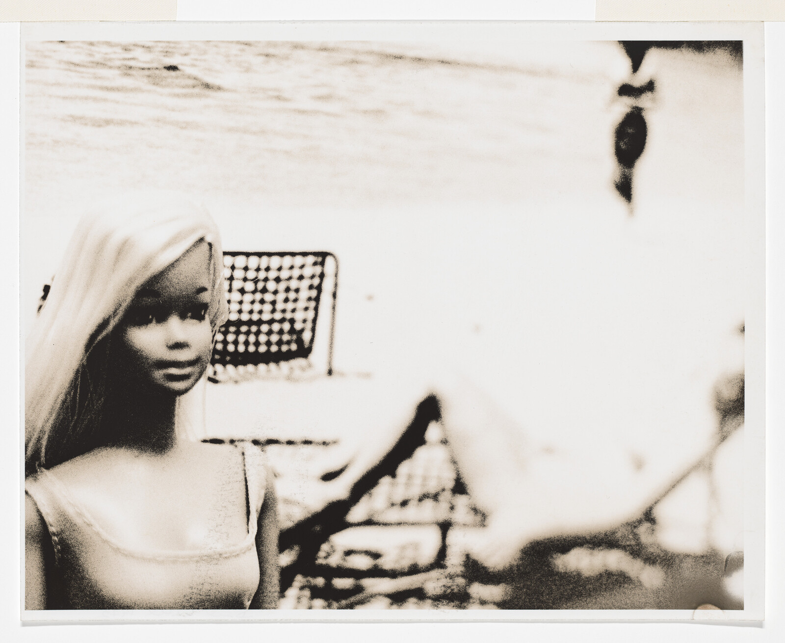 A blonde doll stands on a sandy beach with lounge chairs and a blurred figure behind.