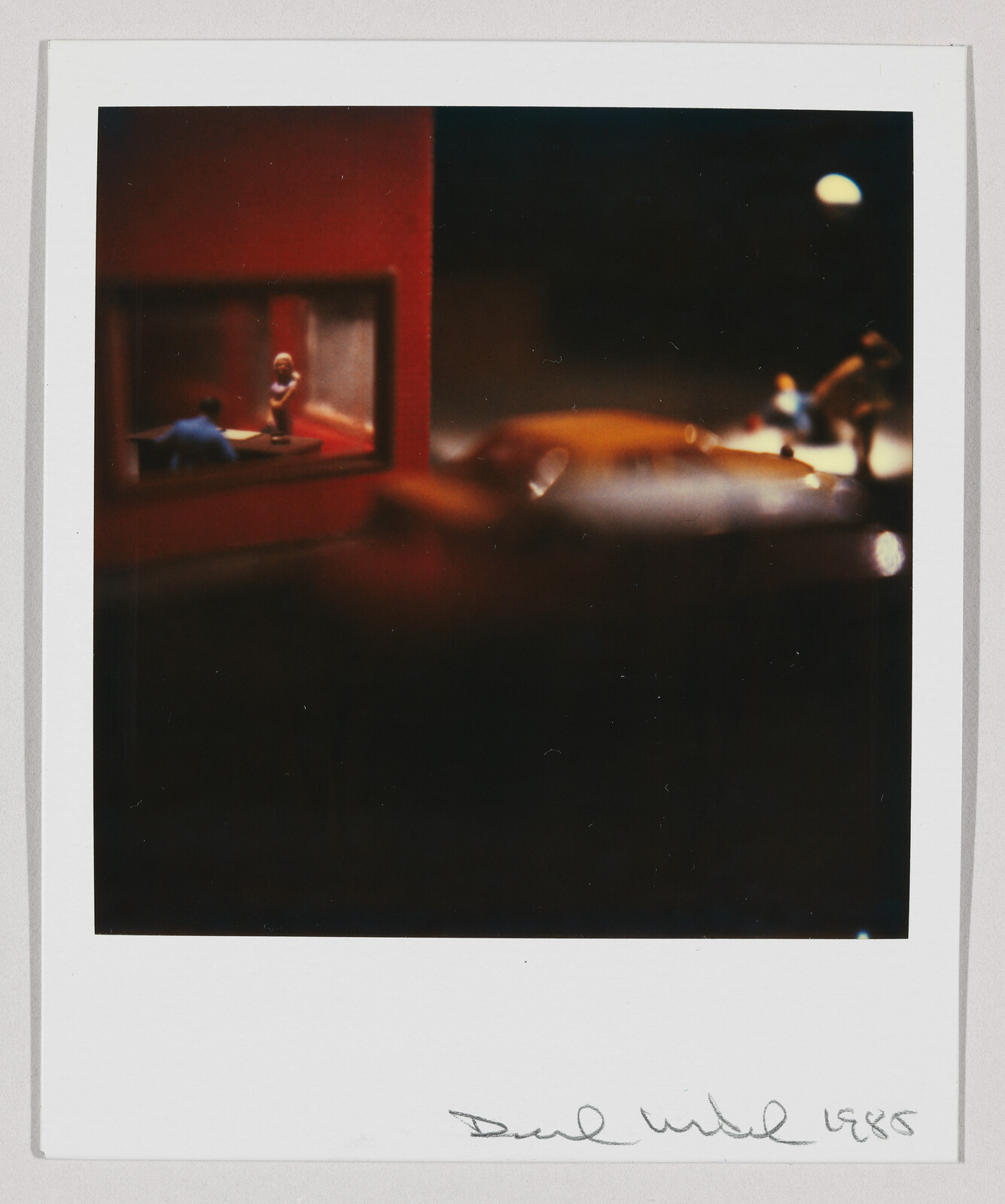 Small lit room with two people talking viewed from a dark street beside a parked car.