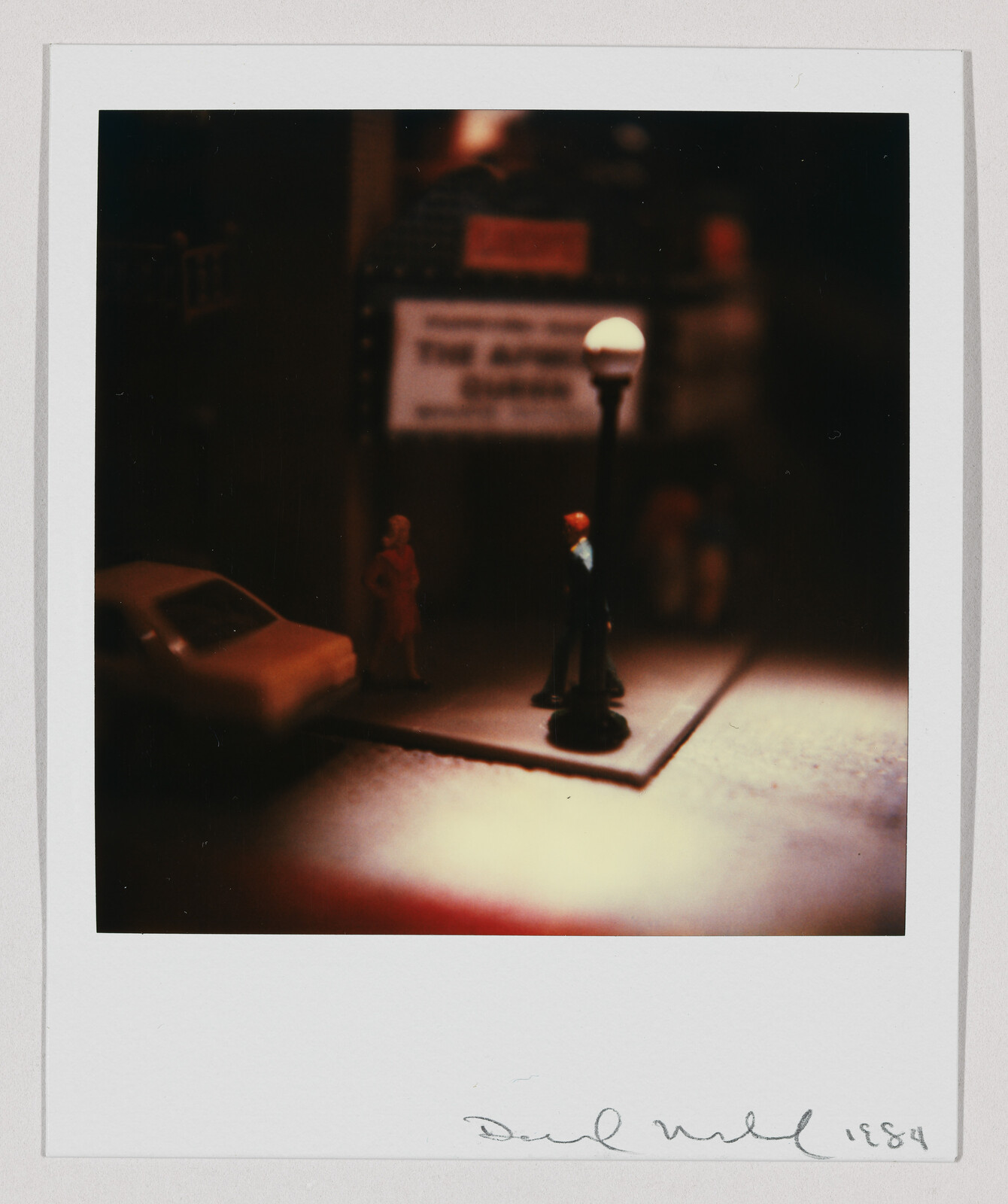 Miniature street scene with two small figures standing under a lit lamppost beside a parked car.