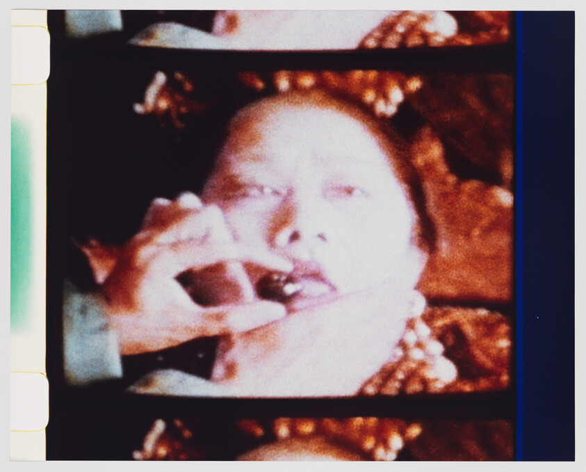 A person wearing a beaded necklace has hands placing a small object into their mouth.