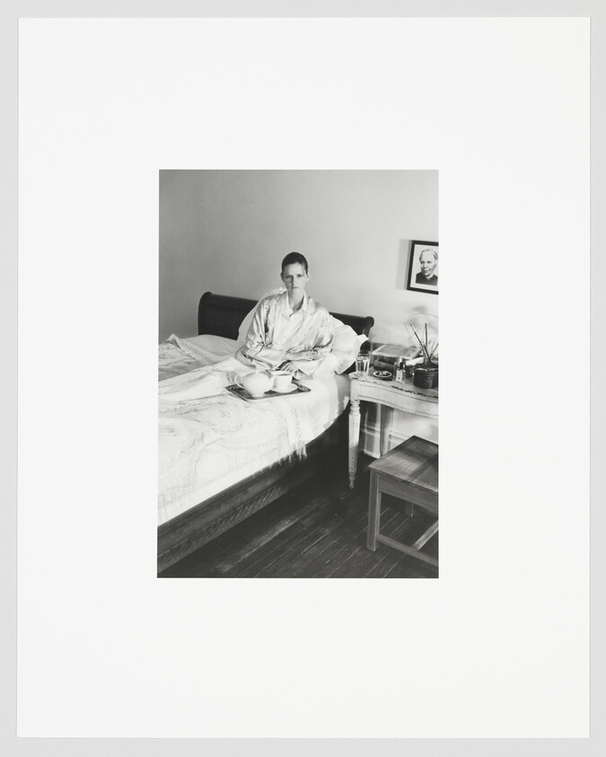 A person sitting on a bed in pajamas with a breakfast tray and a bedside table.