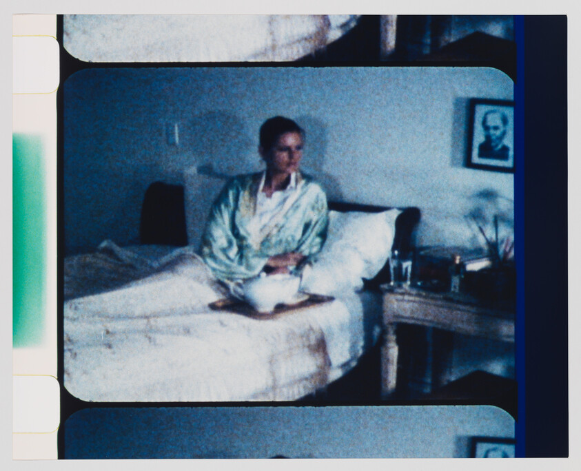 A woman in a robe sits on a bed holding a bowl, with a nightstand and glass nearby.
