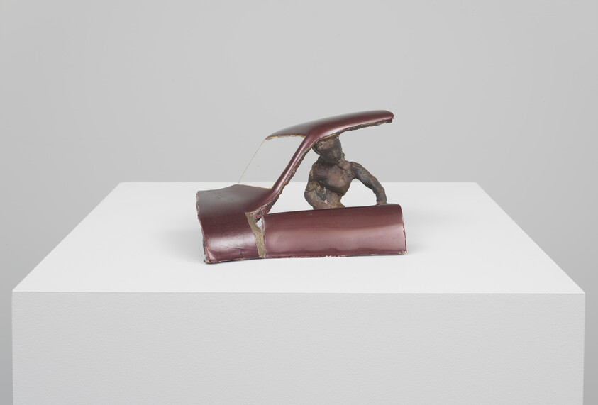Small bronze figure crouches inside a cut maroon car roof displayed on a white pedestal.