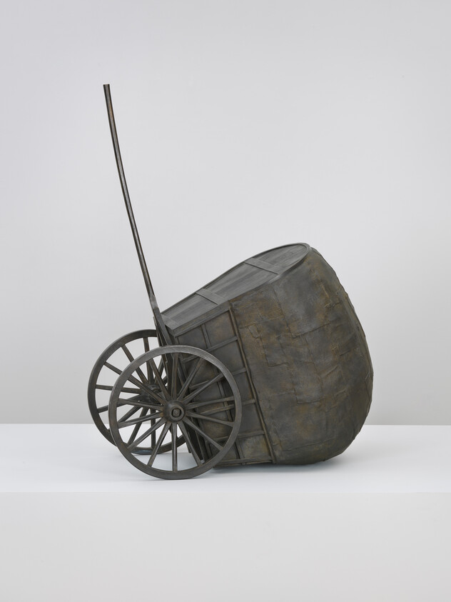 A two-wheeled wooden cart with tall handle is tipped on its side on a white pedestal.
