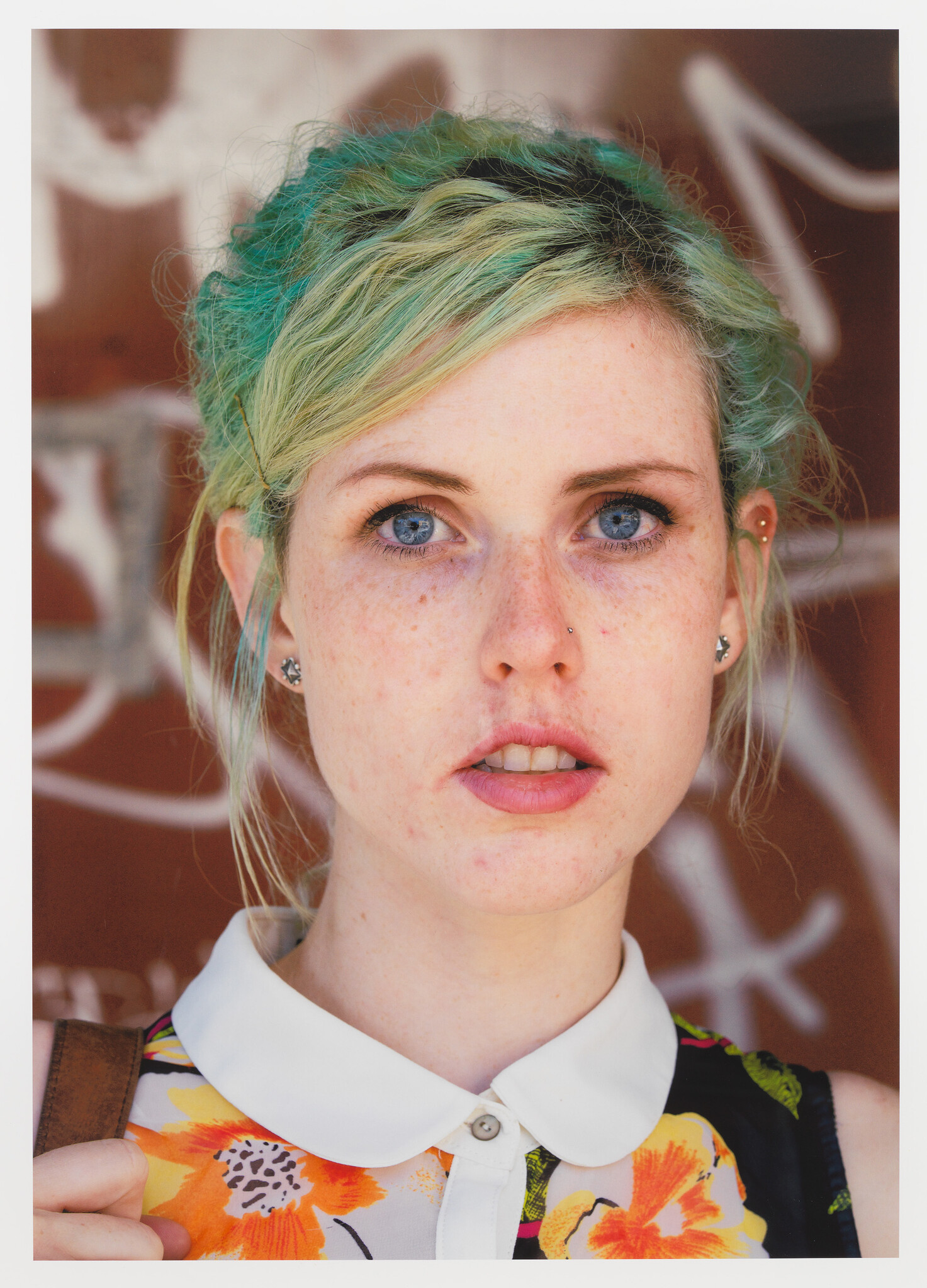 Young woman with green hair and freckles looks directly at the camera against a graffiti background.