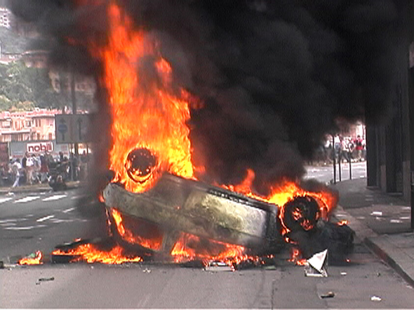 An overturned car engulfed in tall flames and thick black smoke on a city street.