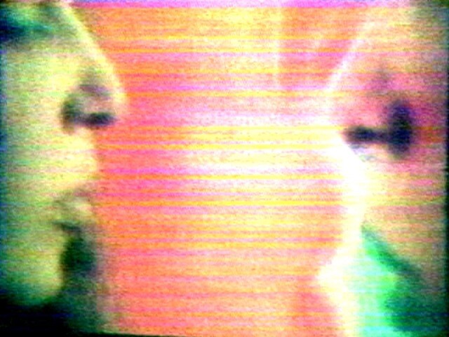Two faces are close together, facing each other, with a colorful, blurry background and visible screen lines.