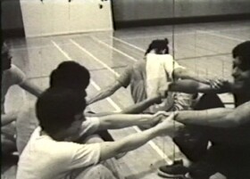 Five people sit on a gym floor in a circle, holding each other's arms and pulling gently.