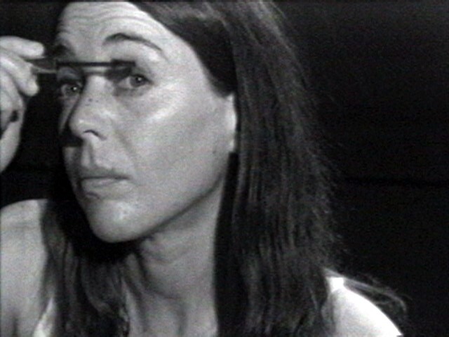 A woman with long dark hair applies mascara to her upper eyelashes, looking slightly upward.