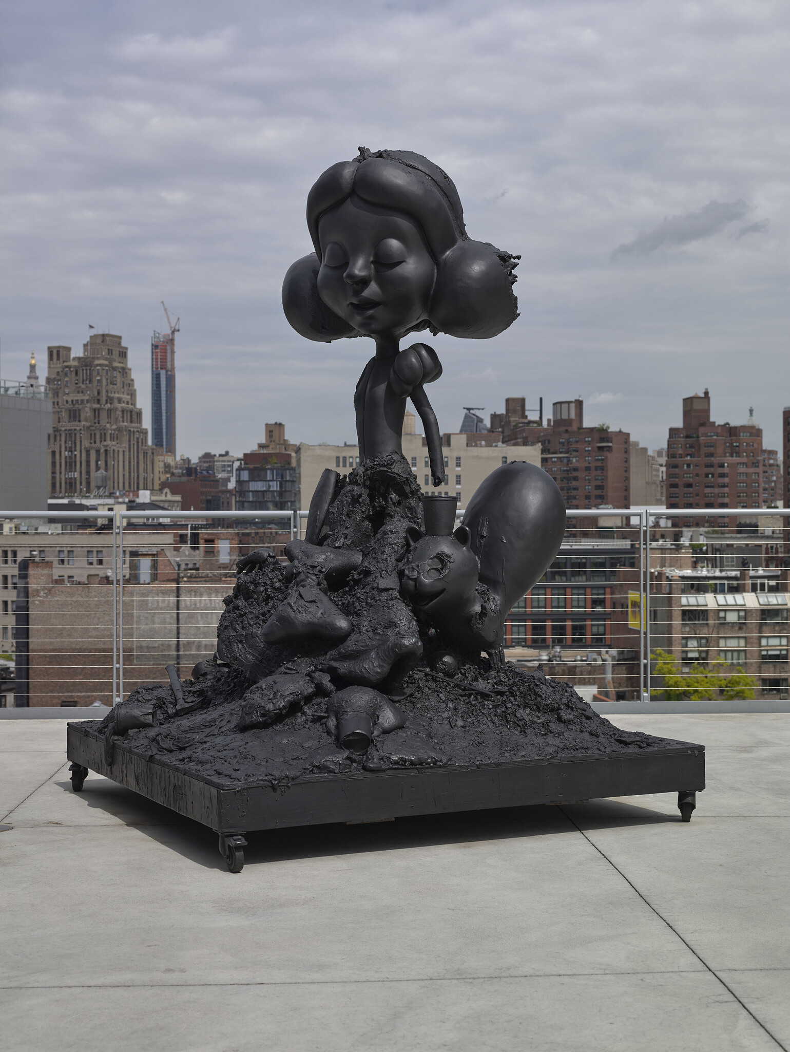 Black melted cartoon-style sculpture of a girl standing on a pile of ruined objects on a rooftop.